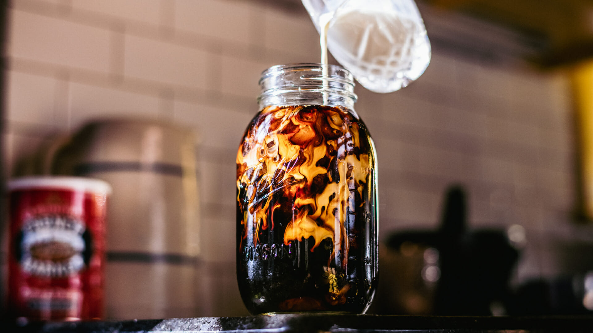 Milk being poured into a glass mason jar of French Market Cold Brew Coffee on a dark kitchen countertop