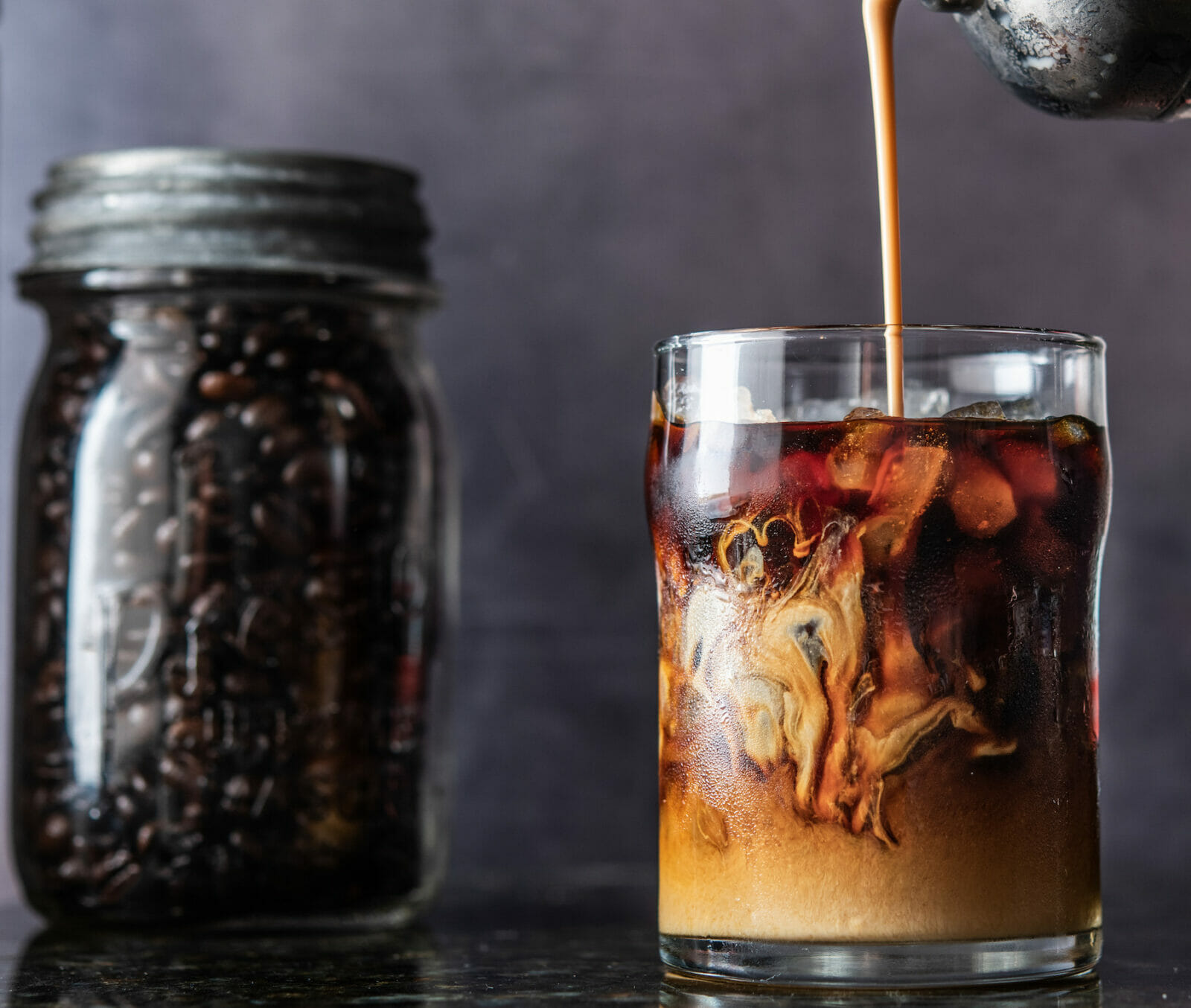 Sweetened condensed milk being poured into glass of dark cold brew coffee with jar of coffee in background