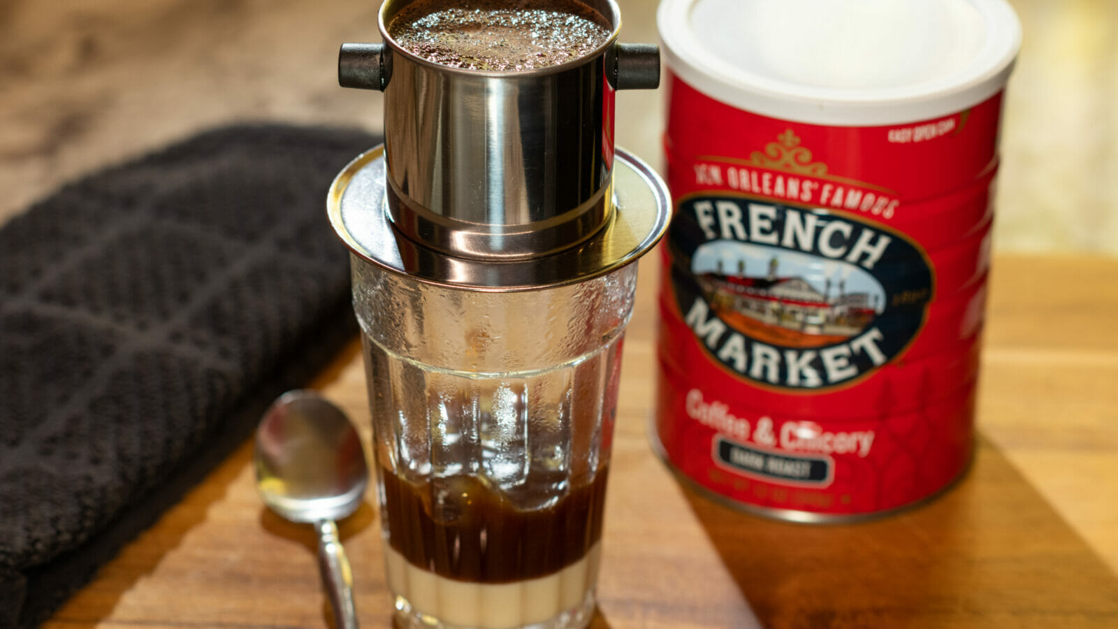 clear glass with condensed milk in bottom and vietnamese coffee filter placed on top with coffee dripping into glass and French Market Coffee can in background