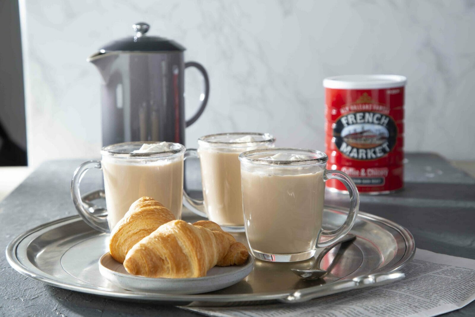 three clear mugs of cafe au lait, or coffee with hot milk, with can of French Market Coffee & Chicory and a metal pitcher of hot milk in the background